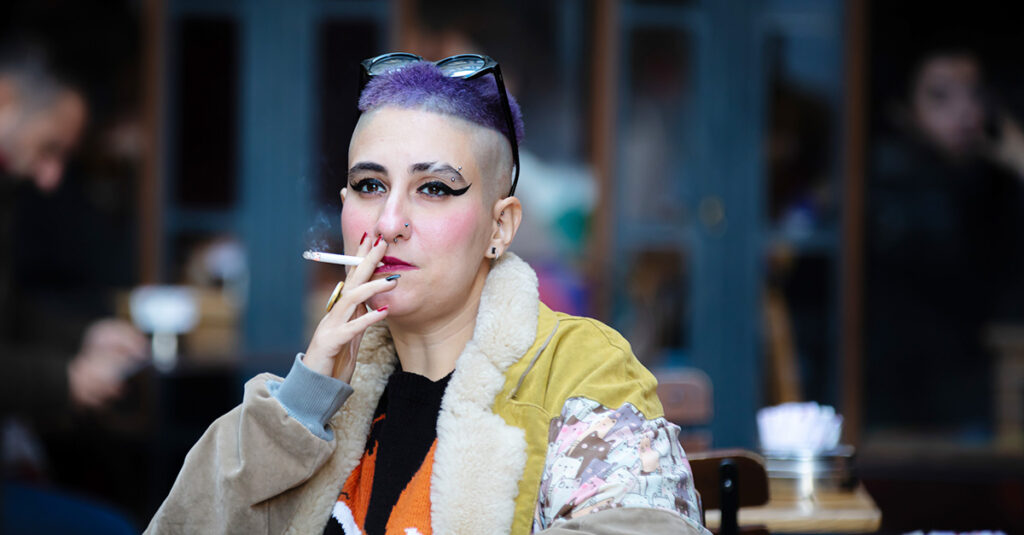 Person with purple hair and bold makeup smoking a cigarette at an outdoor cafe table.