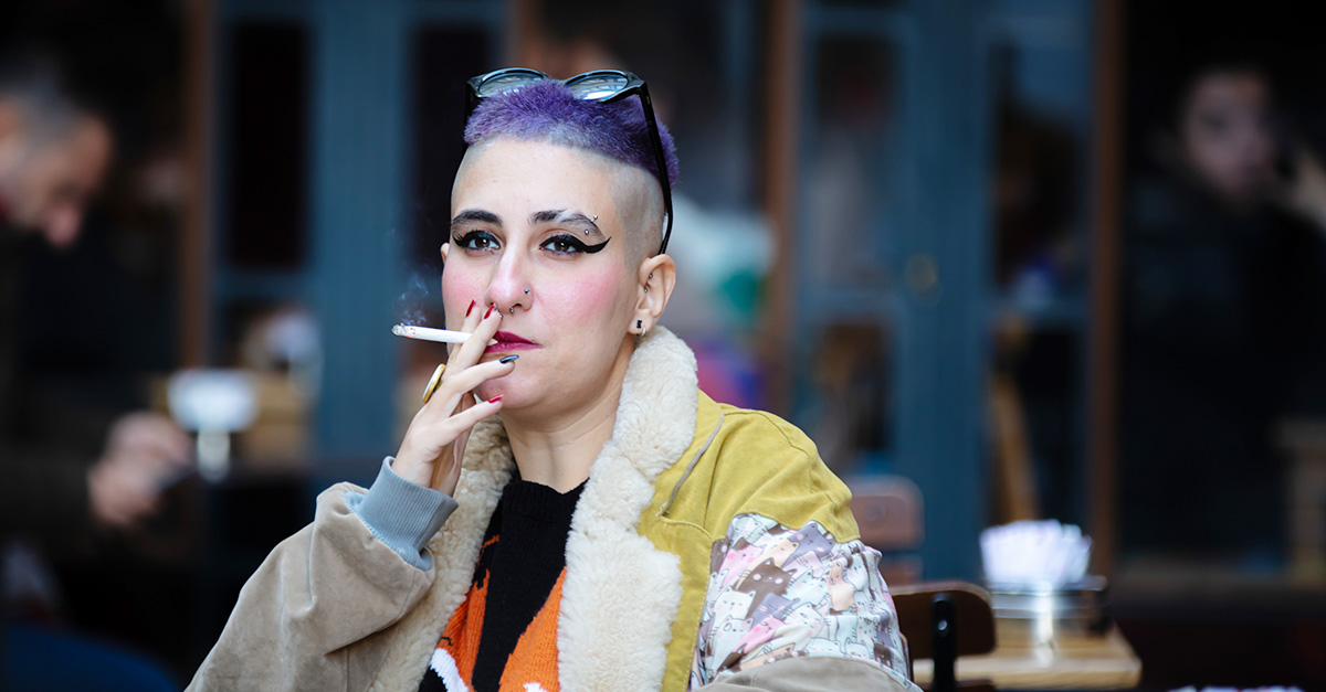 Person with purple hair and bold makeup smoking a cigarette at an outdoor cafe table.